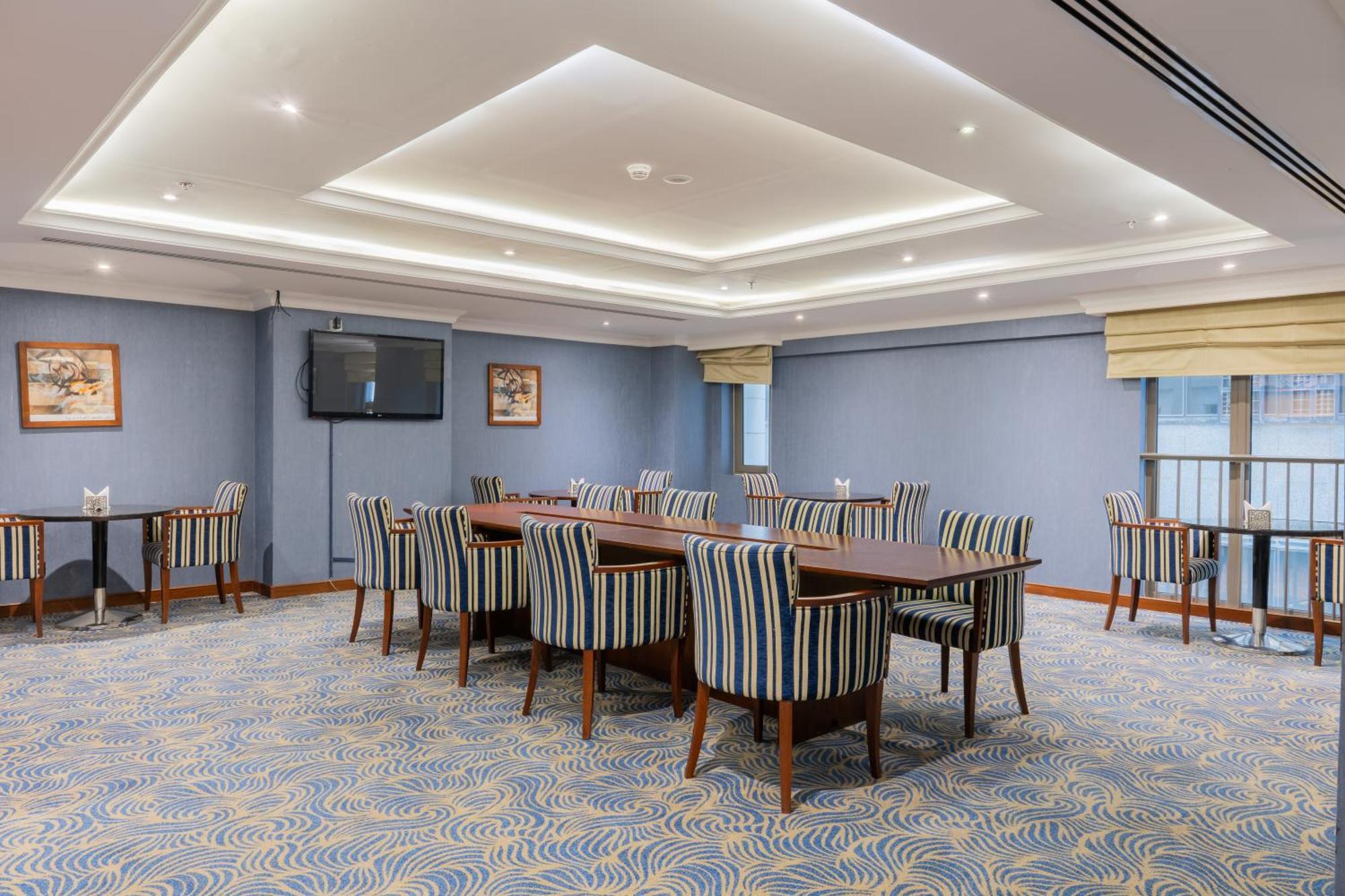 Al Aqeeq Madinah Hotel Medina Exterior foto The photo shows a spacious conference or meeting room. It features a light blue color scheme with a patterned carpet. There are several rectangular tables arranged in a group, surrounded by striped chairs. The room is well-lit with modern ceiling lig