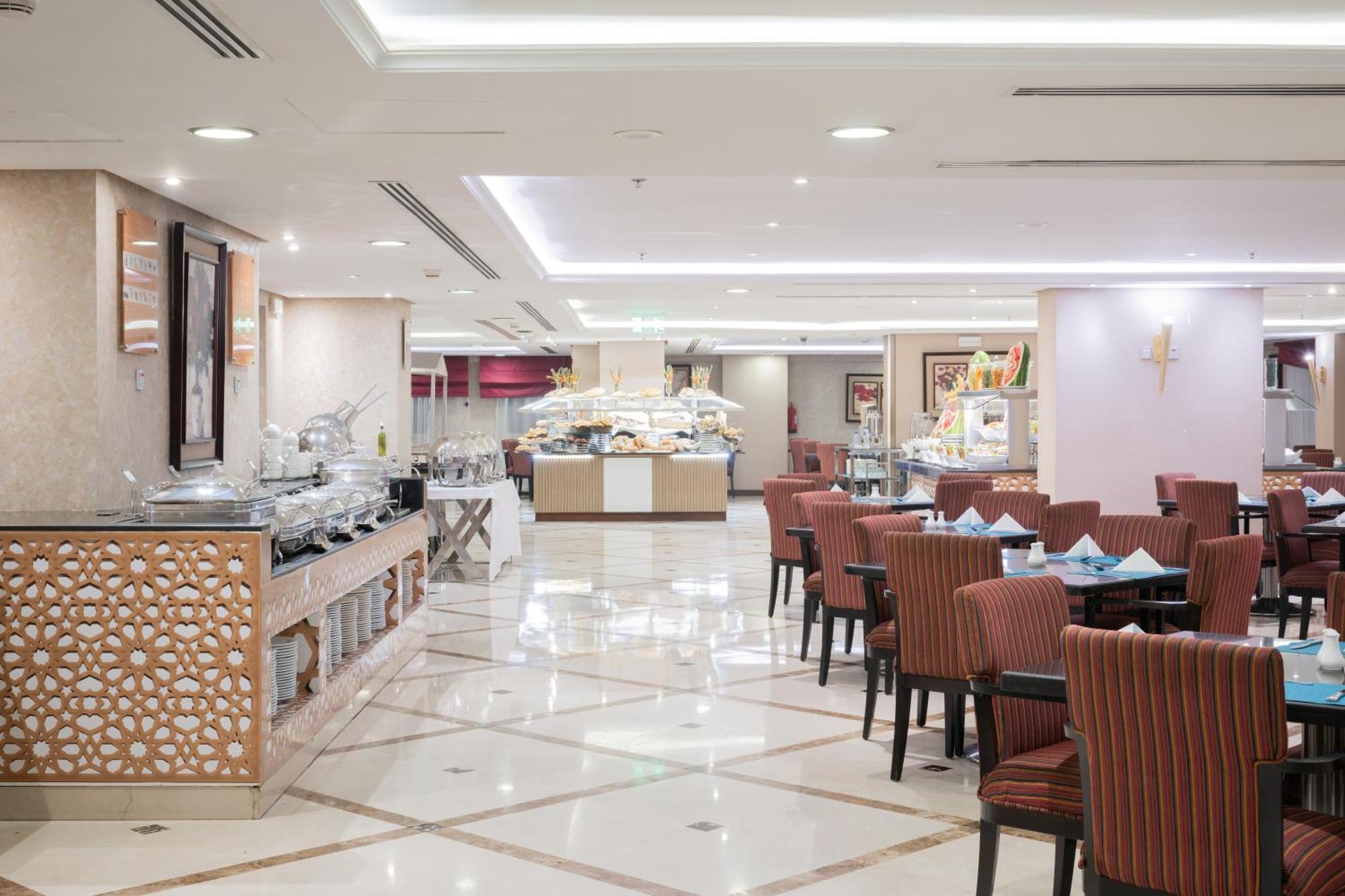 Al Aqeeq Madinah Hotel Medina Exterior foto The photo shows an elegantly designed restaurant or dining area. It features a spacious layout with a glossy tiled floor and bright, soft lighting. There are several dining tables with striped chairs arranged neatly. Along one side, there's a buffet 
