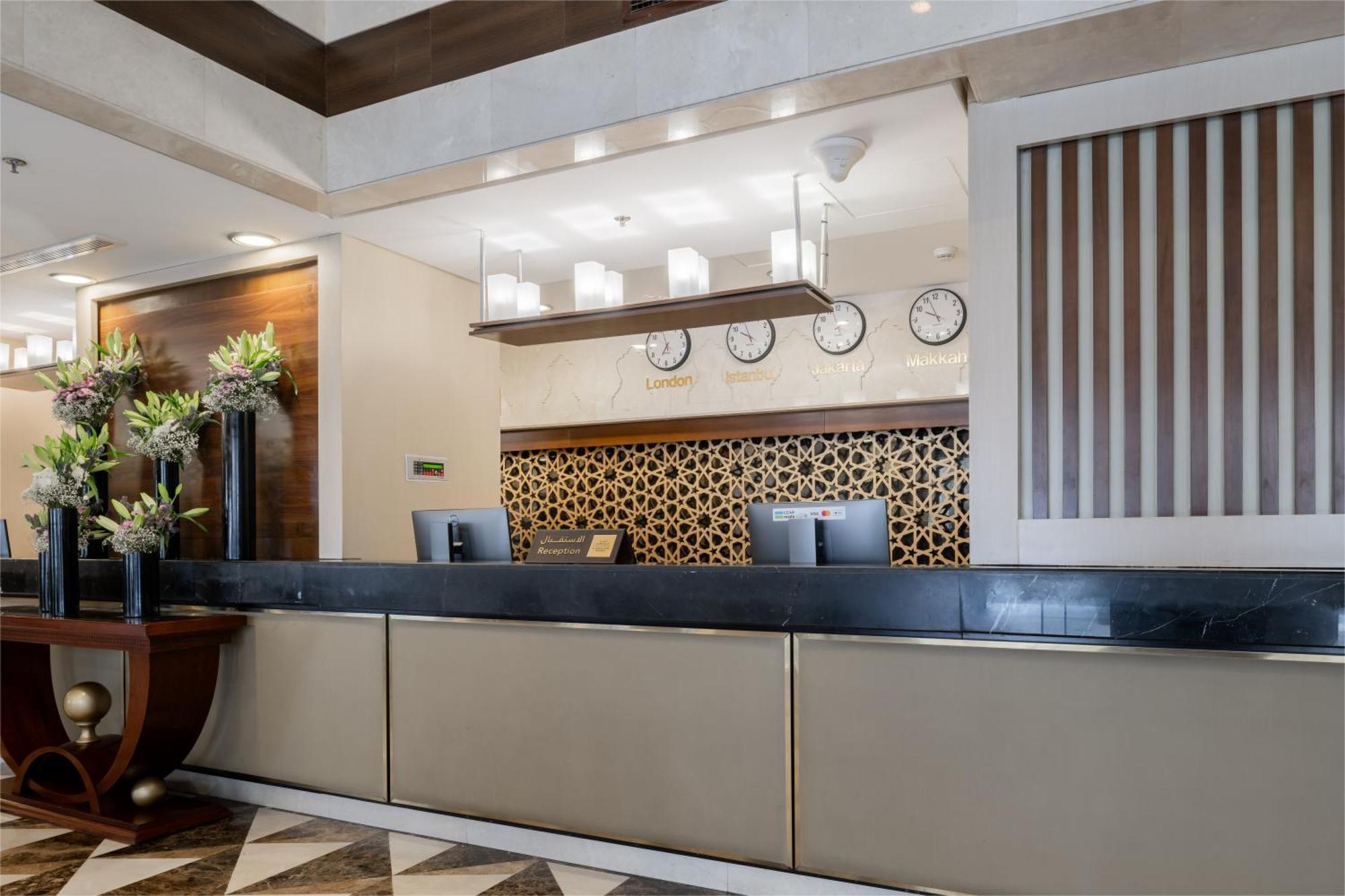 Al Aqeeq Madinah Hotel Medina Exterior foto The photo shows the interior of a hotel reception area. The reception desk is designed with a sleek, modern look featuring a dark countertop and a light-colored front. Above the desk, there are several clocks displaying different times, suggesting an