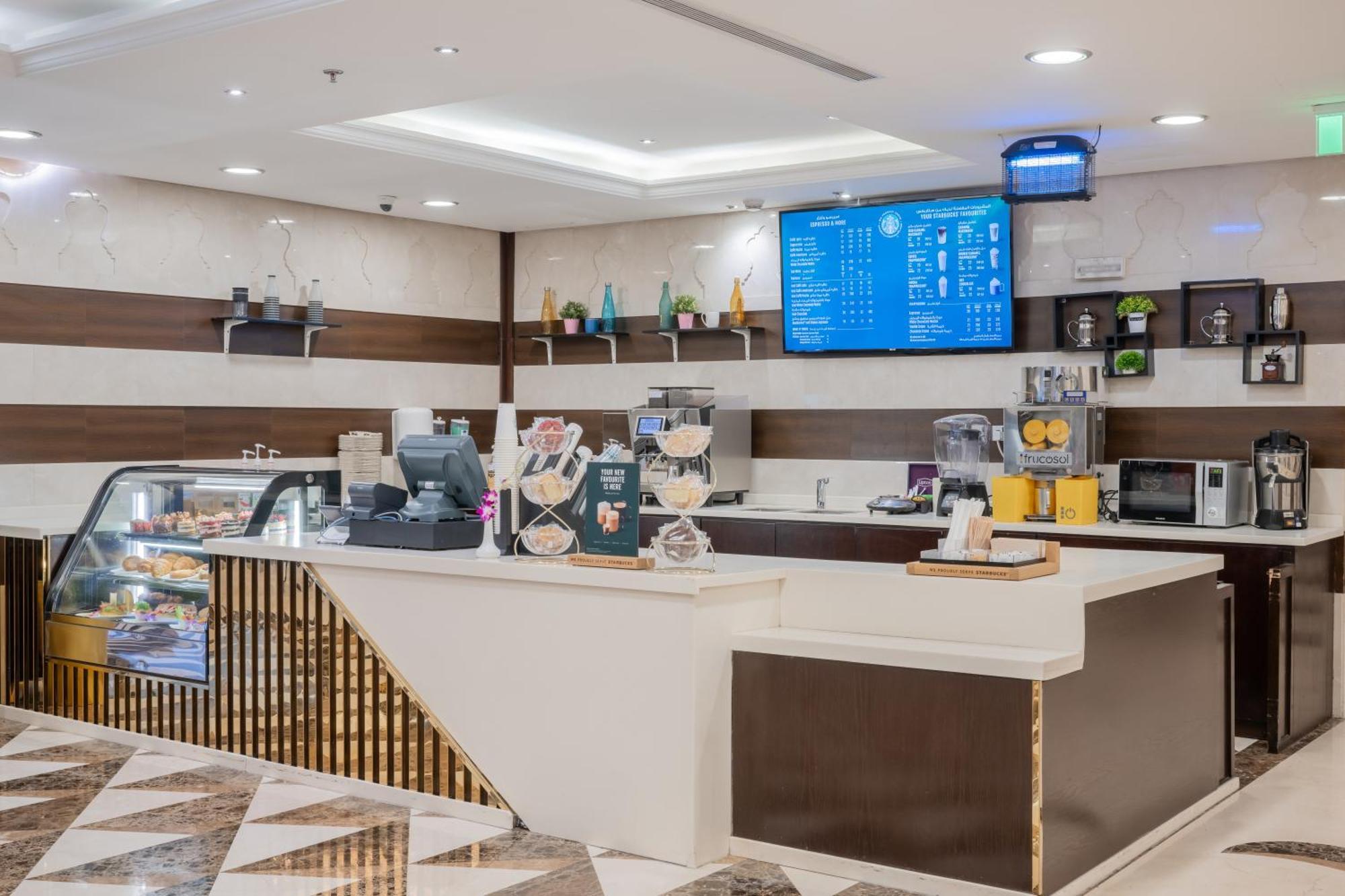 Al Aqeeq Madinah Hotel Medina Exterior foto The photo shows a modern café or coffee shop interior. There's a counter area made of smooth white and dark brown materials, featuring a cash register and display items. Behind the counter, shelves are stocked with various beverage items and equipmen