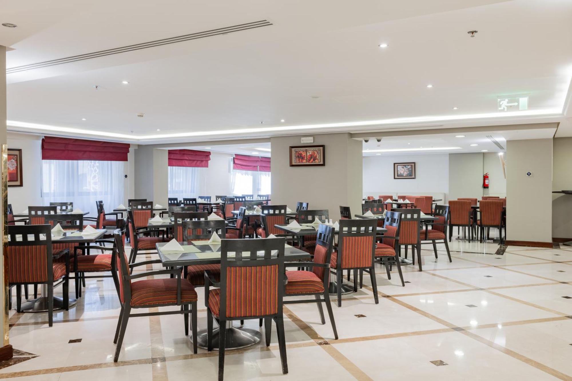 Al Aqeeq Madinah Hotel Medina Exterior foto The photo shows the interior of a restaurant or dining area. The space features several tables arranged with chairs, most of which have striped orange and black upholstery. The flooring is likely marble or tile, and the lighting is bright, creating a