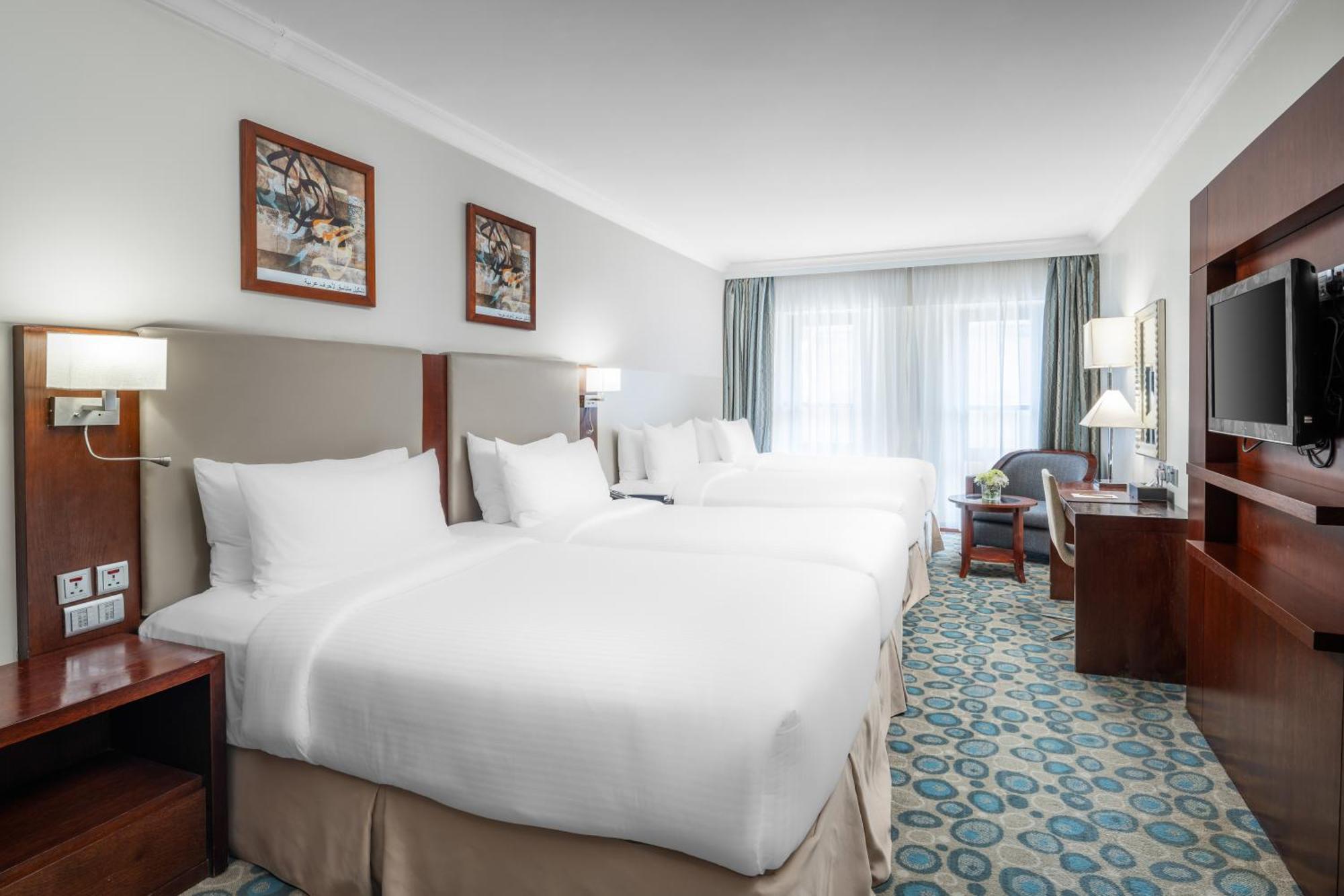 Al Aqeeq Madinah Hotel Medina Exterior foto The photo shows a hotel room featuring a modern, clean design. There are two large beds with white linens positioned side by side. Above the beds are framed artworks, and soft lighting is provided by wall sconces. To the right, there is a wooden ente