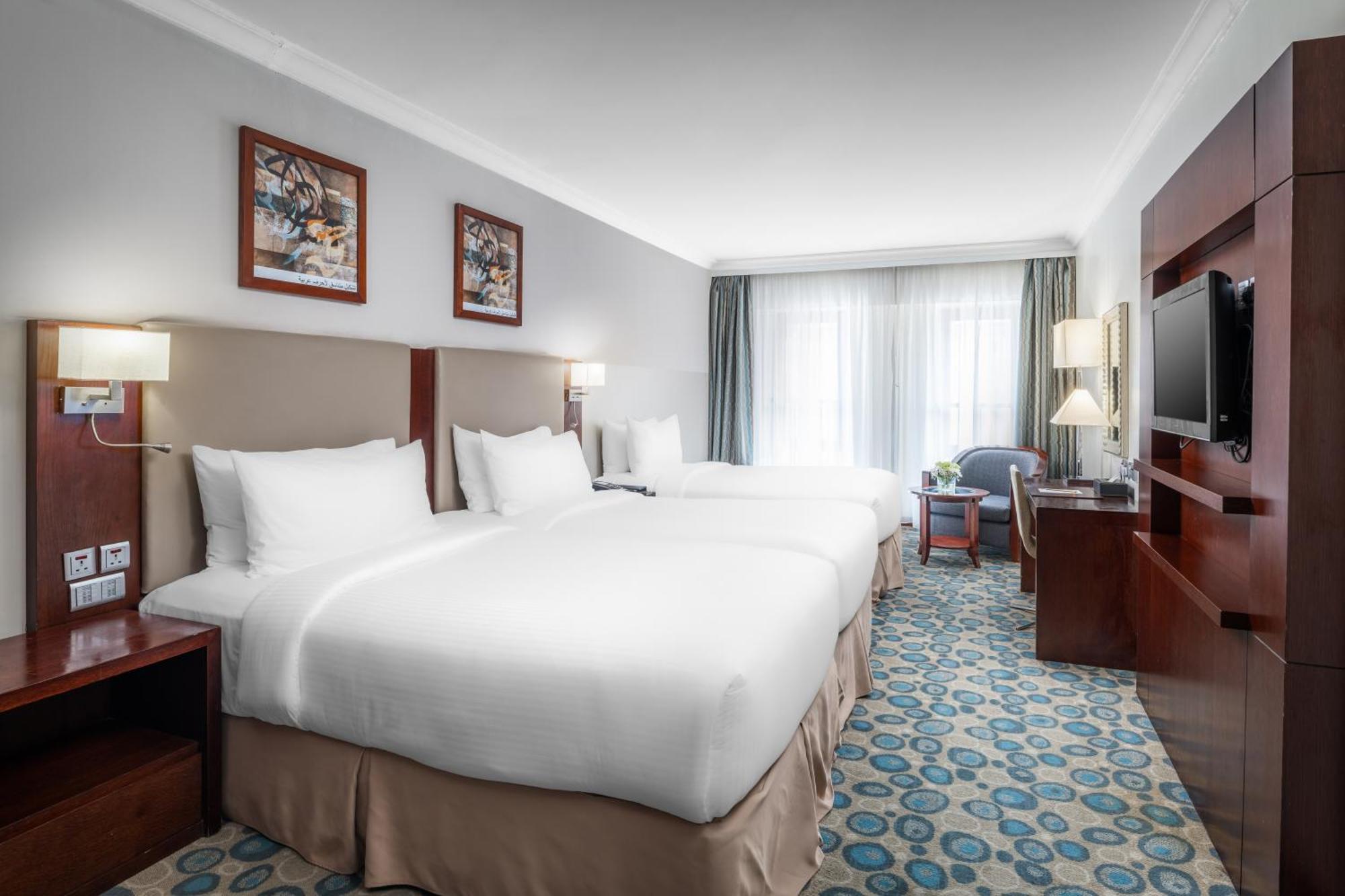 Al Aqeeq Madinah Hotel Medina Exterior foto The photo shows a well-furnished hotel room featuring two large beds with white bedding. The walls are adorned with framed artwork. There are bedside lamps next to each bed, and a flat-screen TV is mounted on the wall. A large window allows natural l