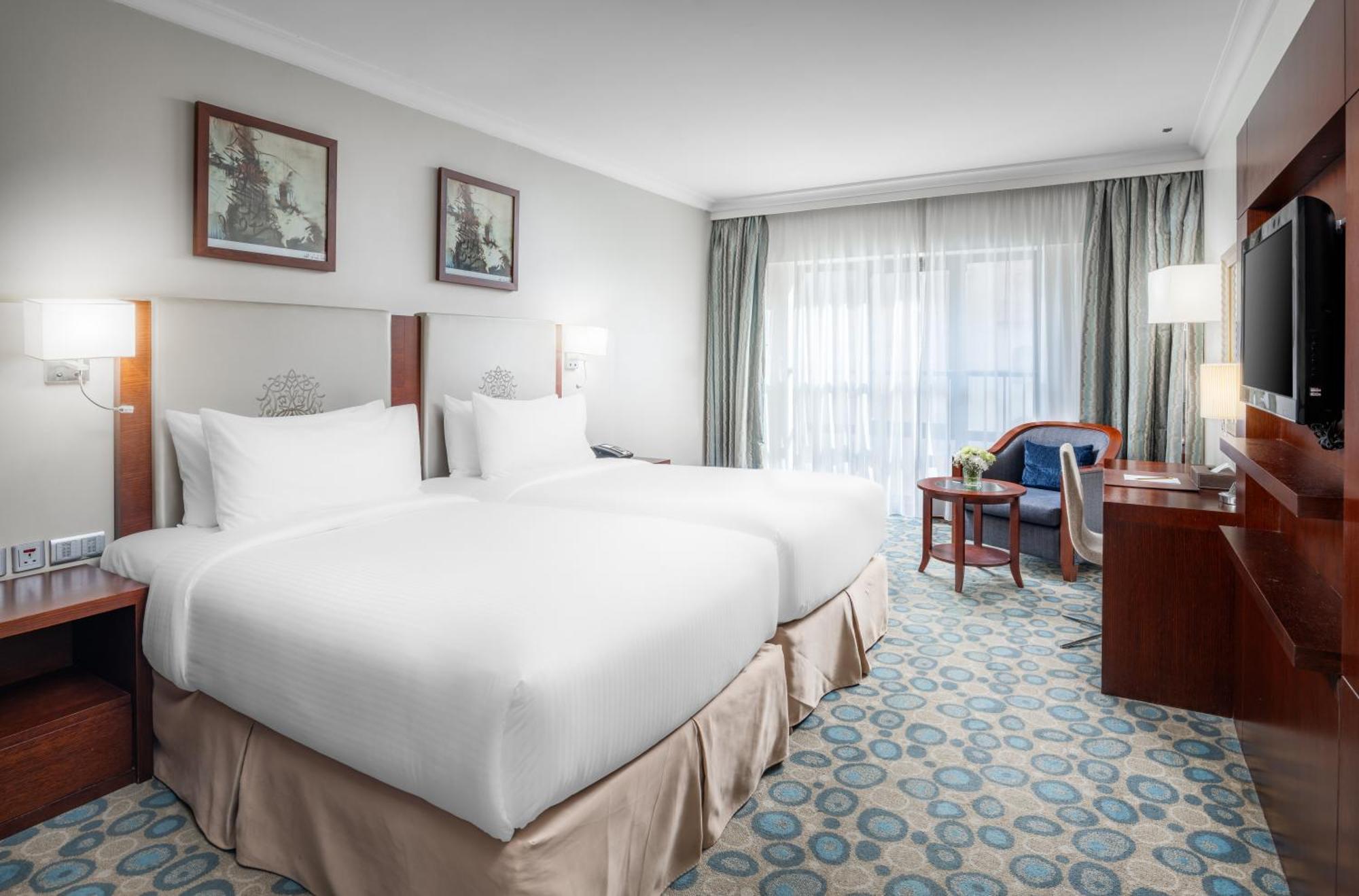 Al Aqeeq Madinah Hotel Medina Exterior foto The photo shows a hotel room featuring two white beds with plush bedding. There are decorative artworks on the walls above the beds. A large window with sheer curtains allows natural light into the space, illuminating the room. The floor is covered w