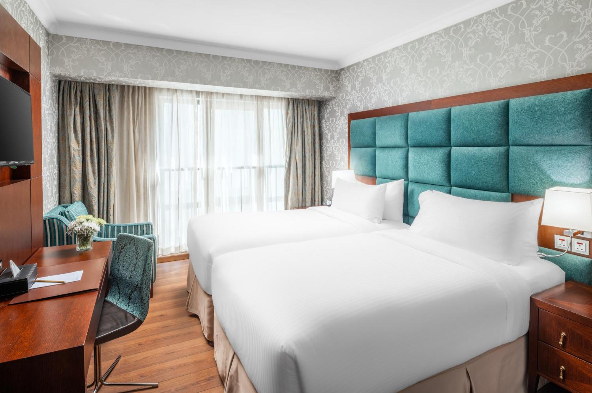Al Aqeeq Madinah Hotel Medina Exterior foto The photo shows a modern hotel room featuring two single beds with white bedding. The headboard is upholstered in a teal fabric. There is a wooden desk and a chair near a window that is covered with sheer curtains and drapes. A cozy armchair in a mat