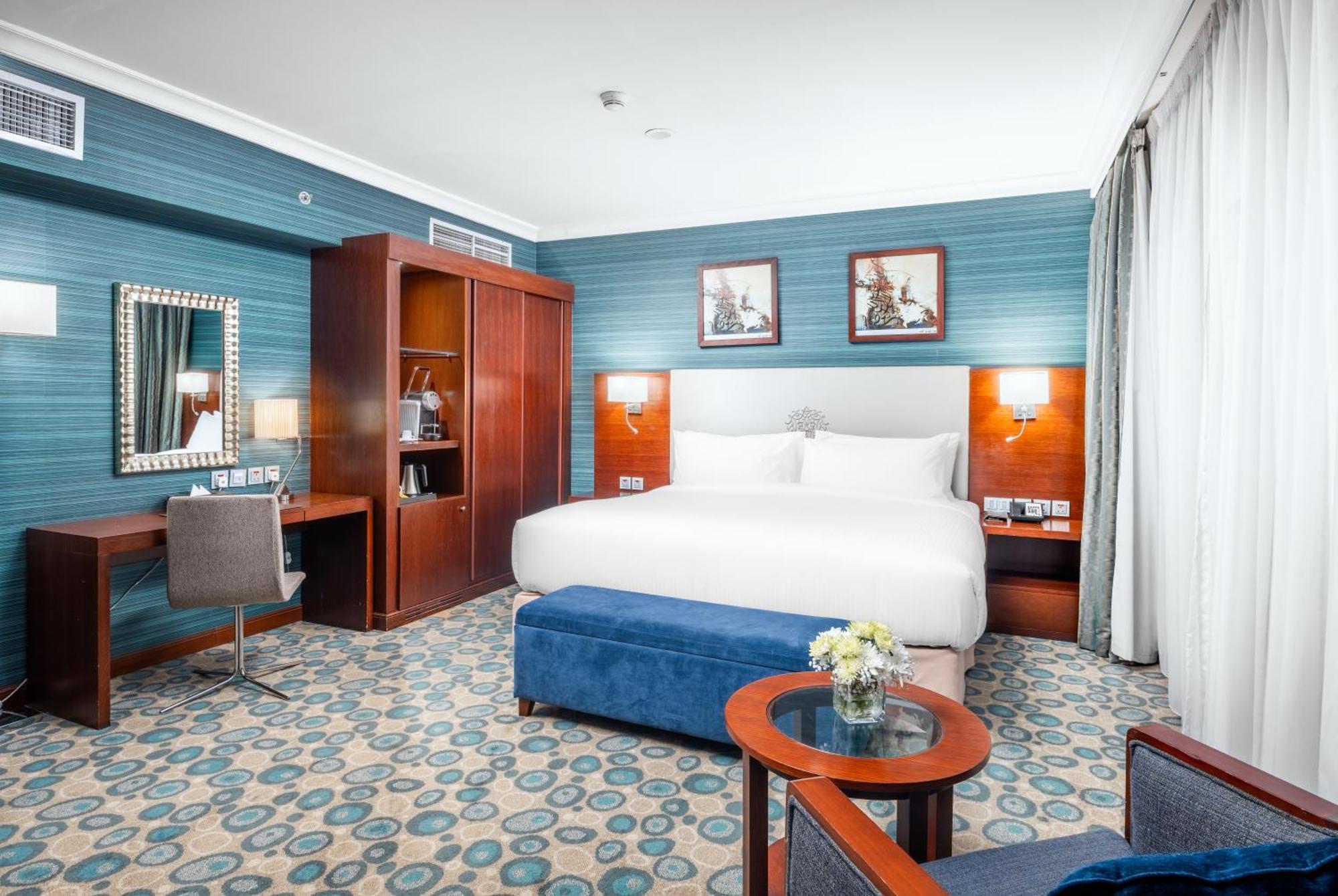 Al Aqeeq Madinah Hotel Medina Exterior foto The photo shows a well-decorated hotel room. It features a large bed with white bedding, flanked by wall-mounted lights. Behind the bed, there are two framed pictures hanging on a blue wall. To the left, there's a wooden wardrobe and a desk with a ch