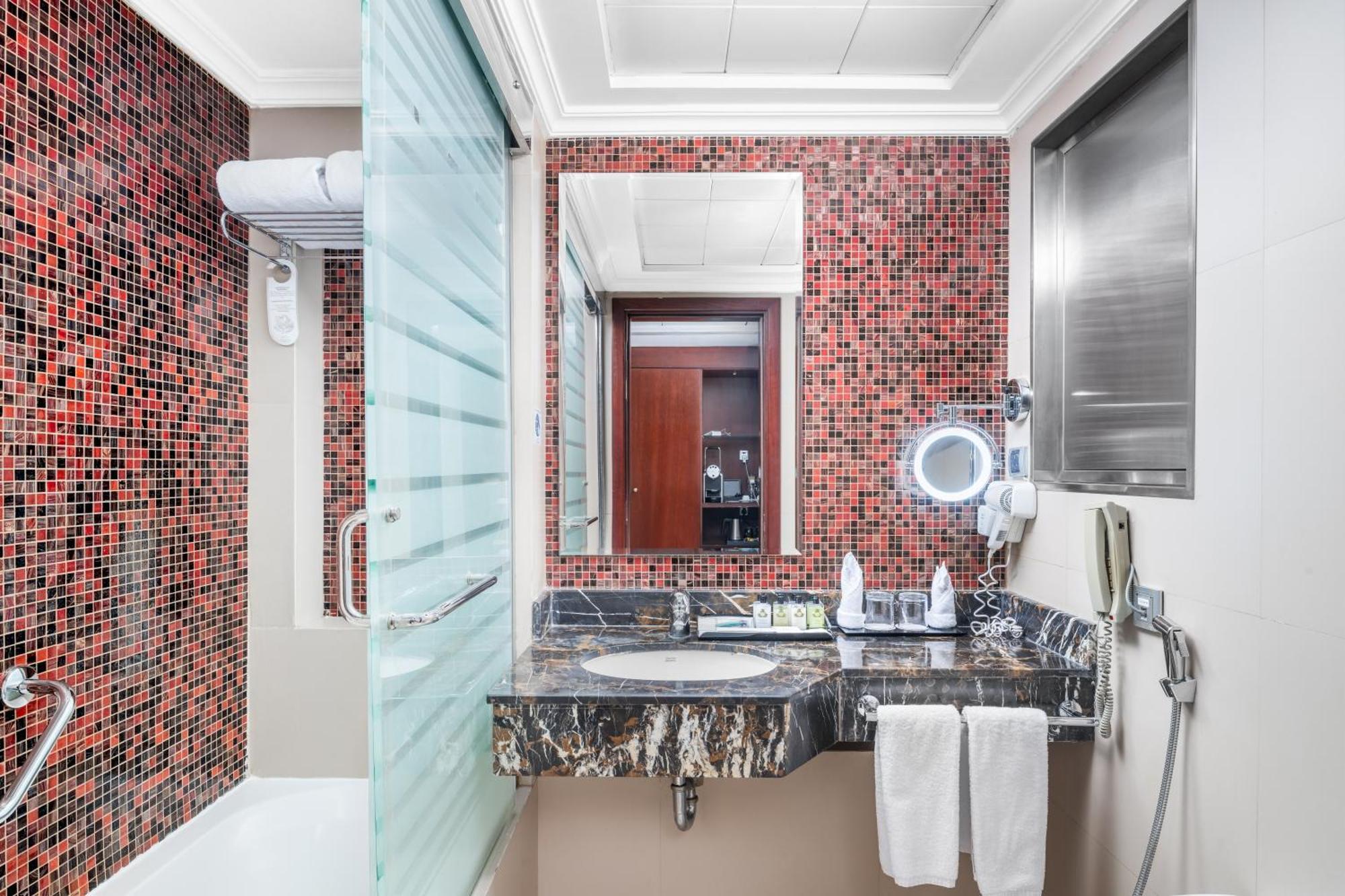 Al Aqeeq Madinah Hotel Medina Exterior foto The photo shows a modern bathroom featuring a mix of design elements. The walls have a vibrant red mosaic tile pattern, adding a bold touch to the space. There is a glass shower enclosure, and next to it, a sleek countertop with a dark, marbled surfa