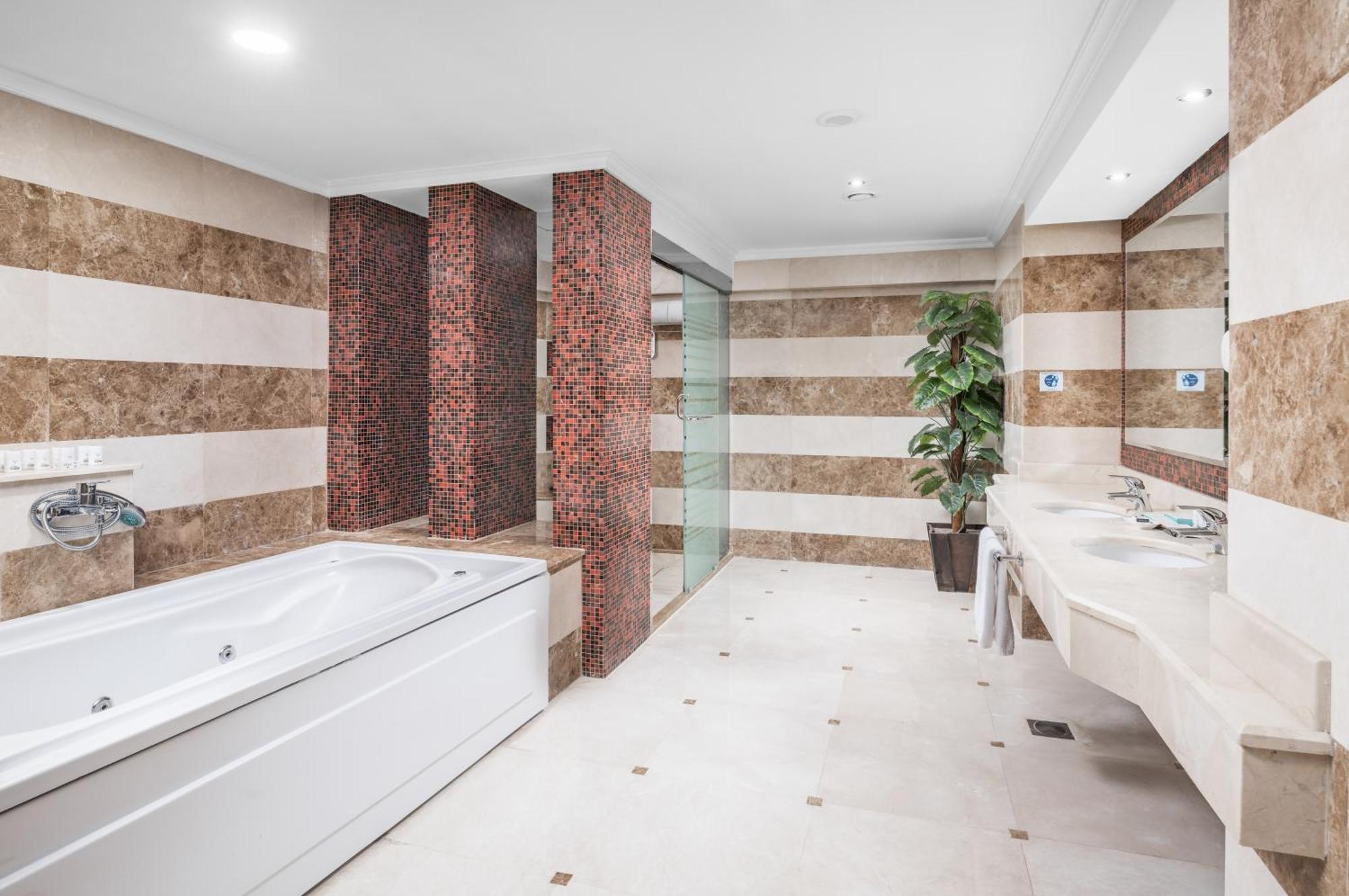 Al Aqeeq Madinah Hotel Medina Exterior foto The photo shows a modern bathroom featuring a spacious layout. On one side, there is a large, white bathtub adjacent to a textured wall with warm, earthy tones. The walls are decorated with alternating horizontal stripes of light and dark colors. 

I