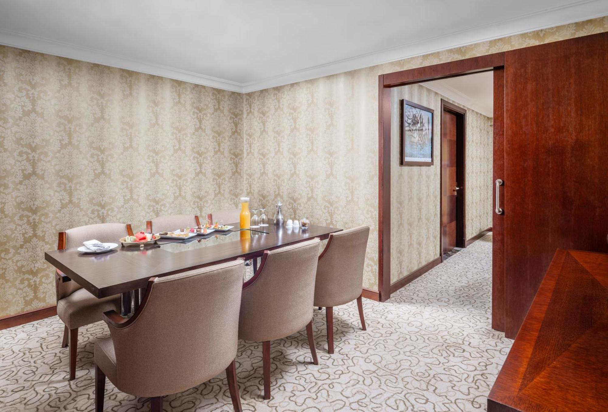Al Aqeeq Madinah Hotel Medina Exterior foto The photo shows a dining room with a stylish and elegant design. There is a large wooden dining table surrounded by plush, beige chairs. On the table, there are various items including a bottle of orange juice, glasses, and other dining accessories. 