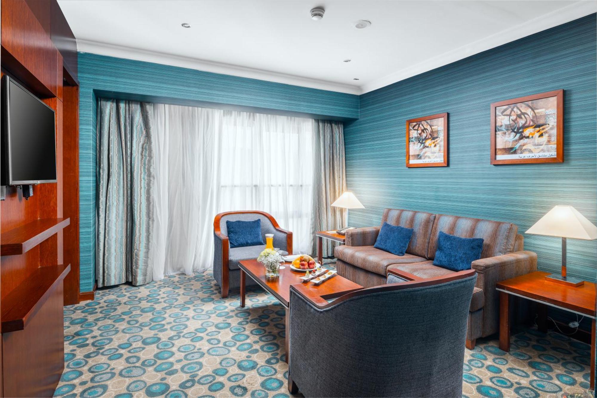 Al Aqeeq Madinah Hotel Medina Exterior foto The photo shows a cozy and modern hotel room featuring a sitting area. The walls are painted a calming teal color, complemented by a patterned carpet in shades of blue and beige. There is a comfortable sofa with cushions, two armchairs, and a wooden 