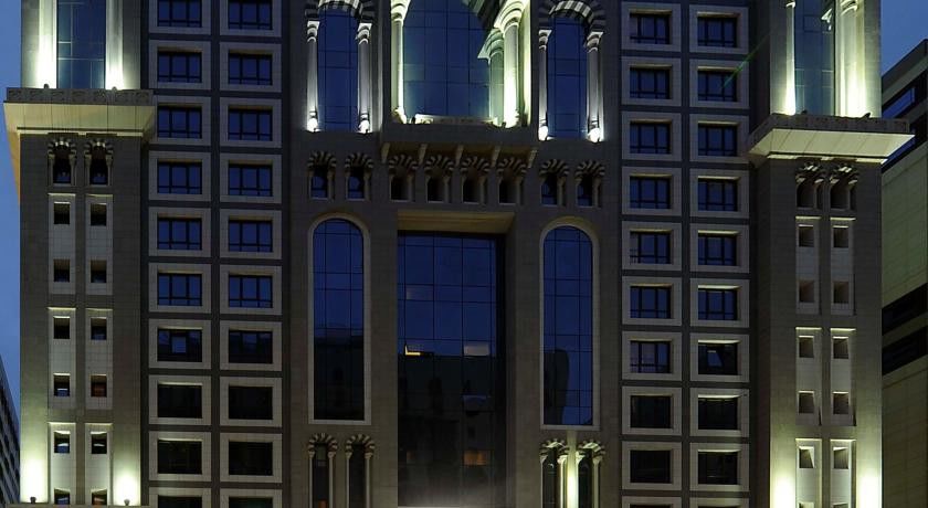 Al Aqeeq Madinah Hotel Medina Exterior foto The image shows the facade of a modern high-rise building illuminated at night. The design features large glass windows arranged in a grid pattern, along with decorative architectural elements at the top, including arches and ornate detailing. The co