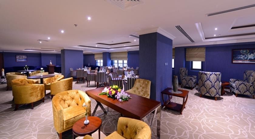 Al Aqeeq Madinah Hotel Medina Exterior foto The photo shows a stylish interior of a restaurant or dining area. It features elegantly arranged seating with plush, gold-colored chairs and a wooden table at the center, adorned with a floral arrangement. The background includes more dining tables,