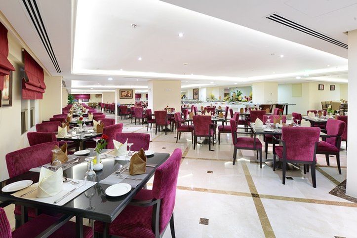 Al Aqeeq Madinah Hotel Medina Exterior foto The photo shows an elegant restaurant interior with a spacious layout. The dining area features multiple tables, each set with white tablecloths and dinnerware. The chairs are upholstered in a rich purple fabric, adding a touch of luxury to the atmos