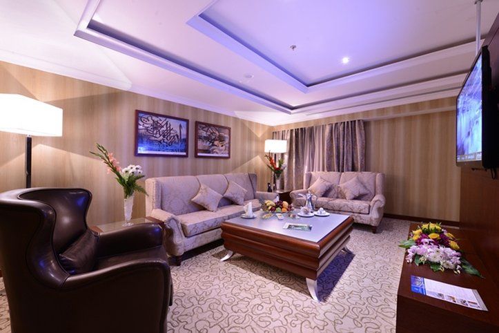 Al Aqeeq Madinah Hotel Medina Exterior foto The photo shows a stylish and comfortable living room. It features two light-colored sofas with decorative cushions arranged around a modern coffee table. In one corner, there's a dark, leather armchair. The walls are adorned with framed artwork, and