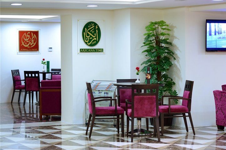 Al Aqeeq Madinah Hotel Medina Exterior foto The photo shows an interior space, likely a dining or lounge area. There are several tables with four chairs each, featuring a modern design with dark wood and plush maroon upholstery. On the walls, there are decorative signs, one of which has Arabic