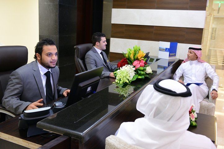 Al Aqeeq Madinah Hotel Medina Exterior foto The photo depicts a reception area with three individuals. Two men are seated behind a reception desk; they are dressed in formal suits and appear to be engaged in conversation, possibly assisting a guest. The desk has a computer and a vibrant arrang